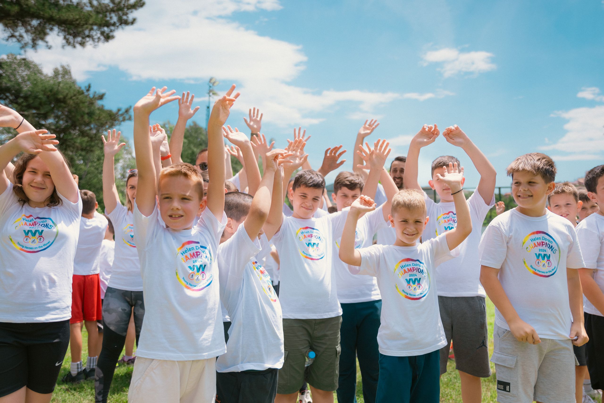 Wycieczki szkolne 1 A group of cheerful children in white T-shirts raising hands in a park during summer.