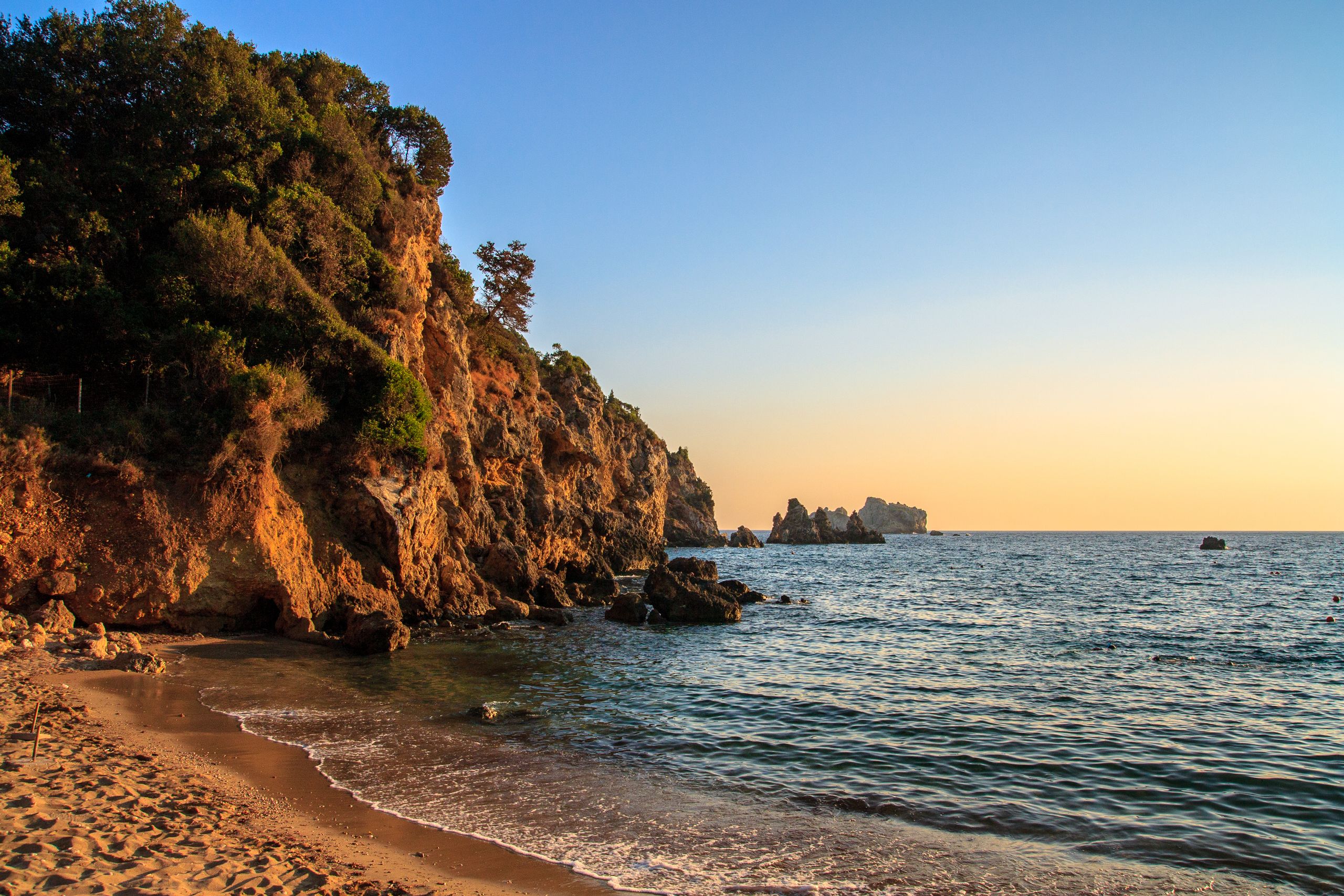 Wczasy i Wycieczki 5 Dramatic sunset over rocky cliffs and tranquil sea on a beach in Corfu, Greece.